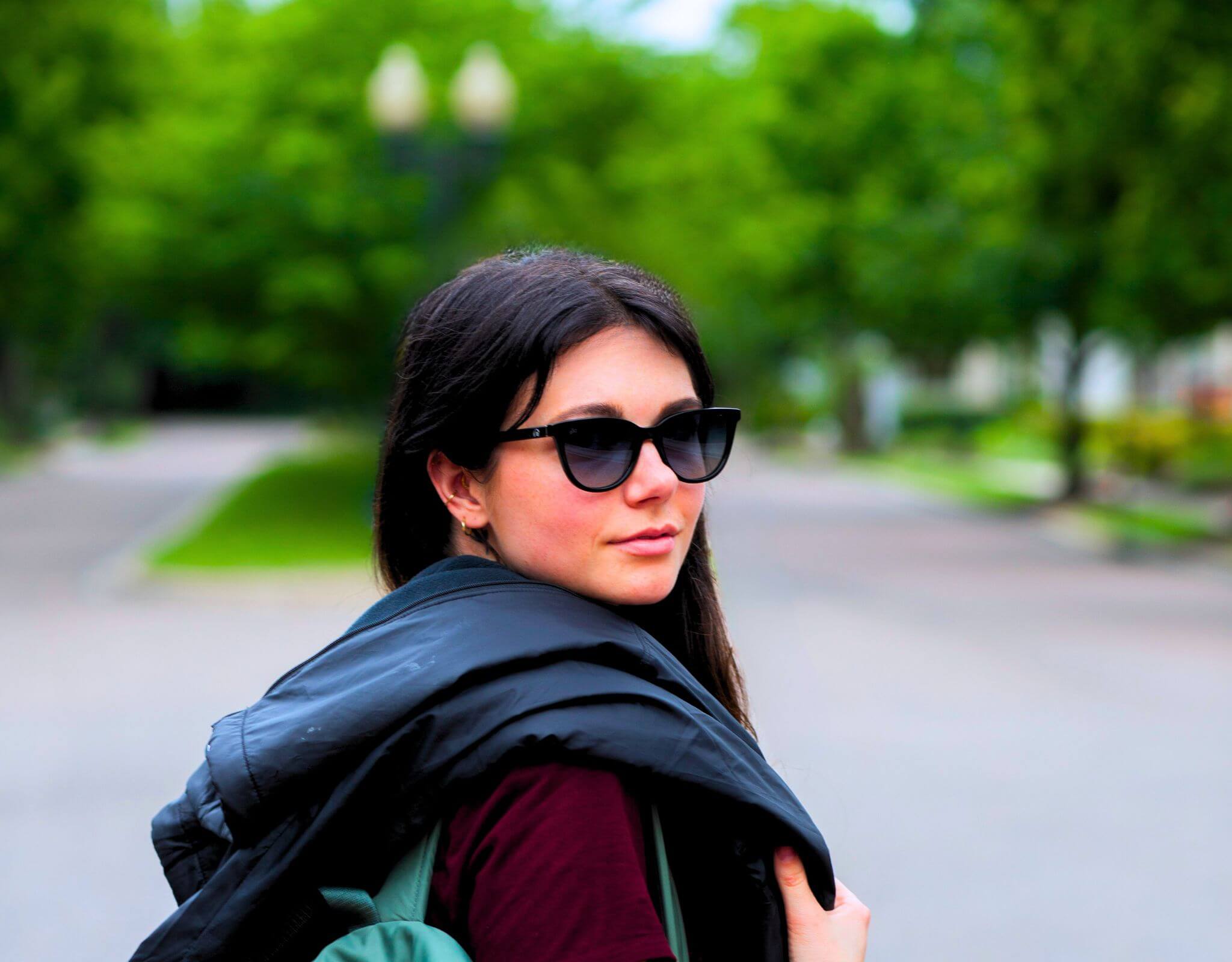 Woman wearing black blue Half Moon Bay cat eye sunglasses for smaller face while walking down a tree-lined street.