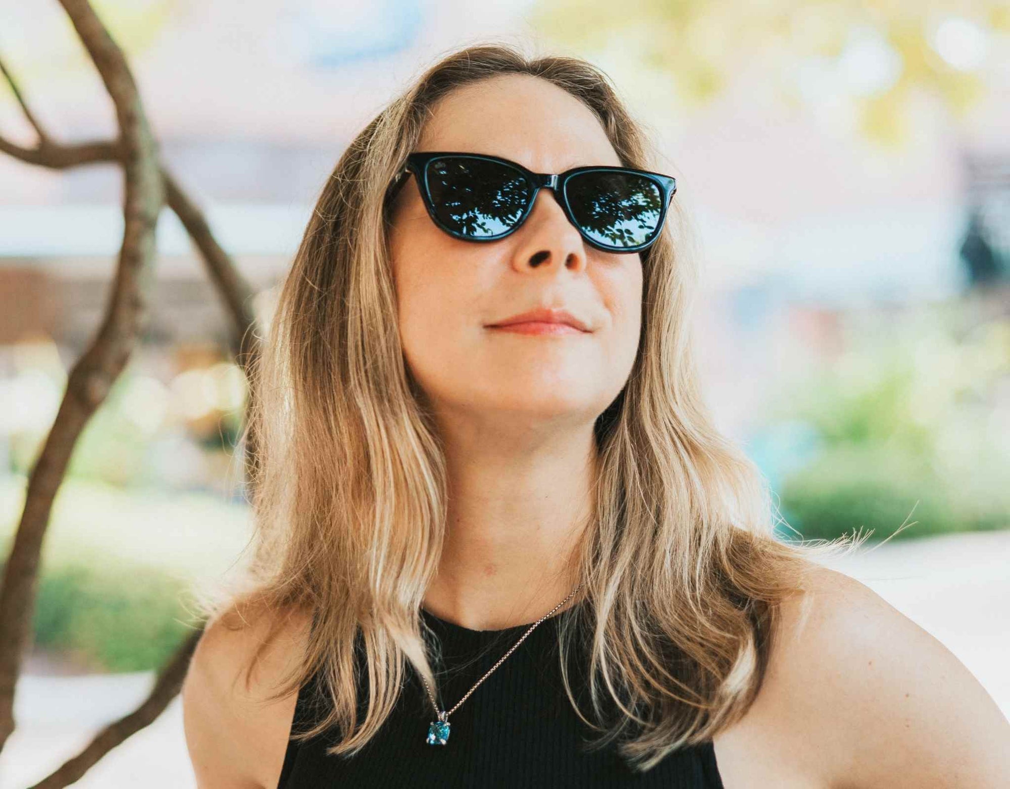 Woman wearing black and blue Half Moon Bay cat eye sunglasses for a smaller face, standing next to a tree.