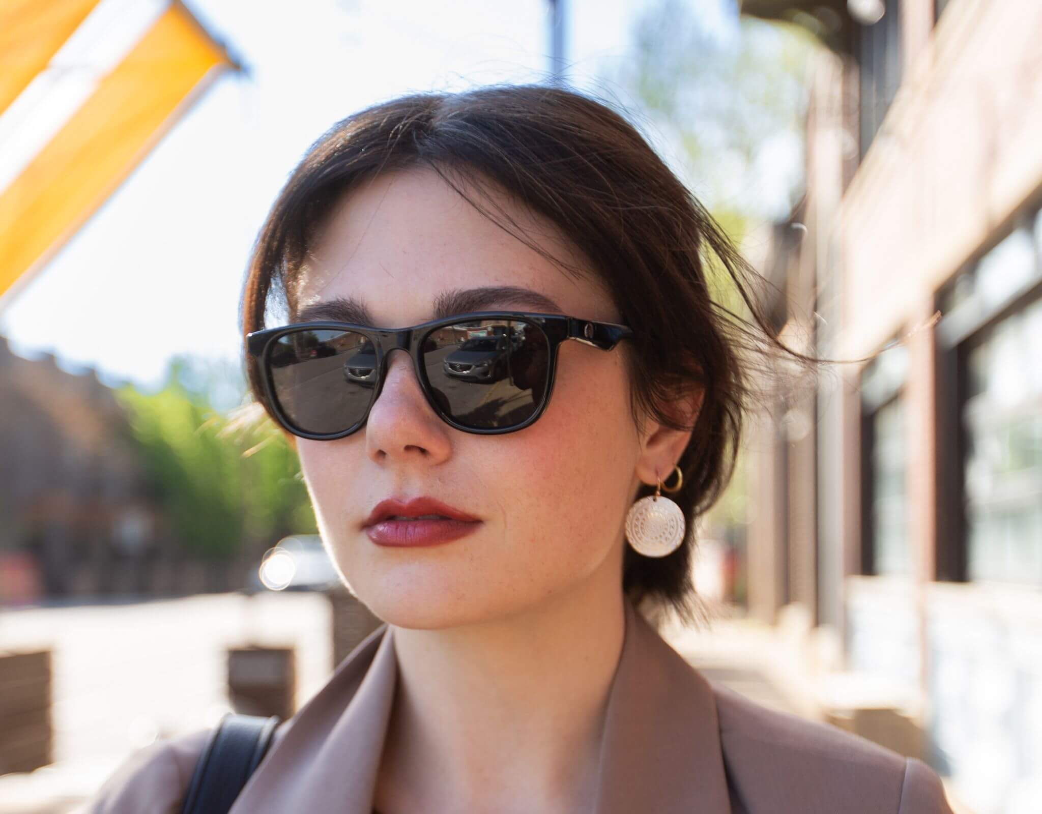 Woman in black grey Outer Banks sunglasses for small faces, styled in a sleek brown suit jacket and earrings on city street