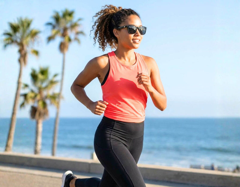 Sunglasses for small faces Outer Banks in black frame with grey lenses worn by a woman with a petite face and small head running along a sunny beach path with ello sunglasses