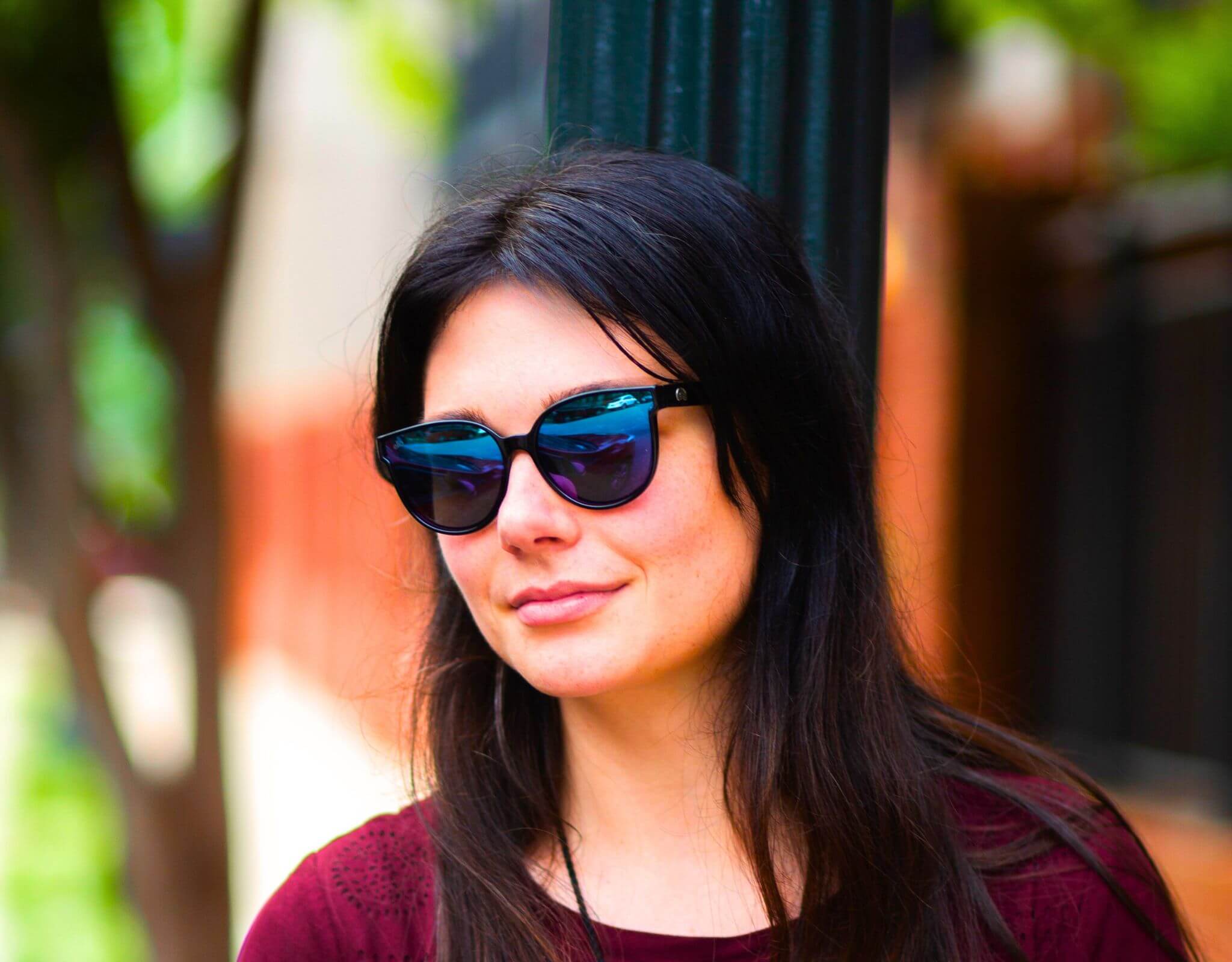 Smiling woman wearing black purple South Beach shades for small faces while leaning against a lamp post.