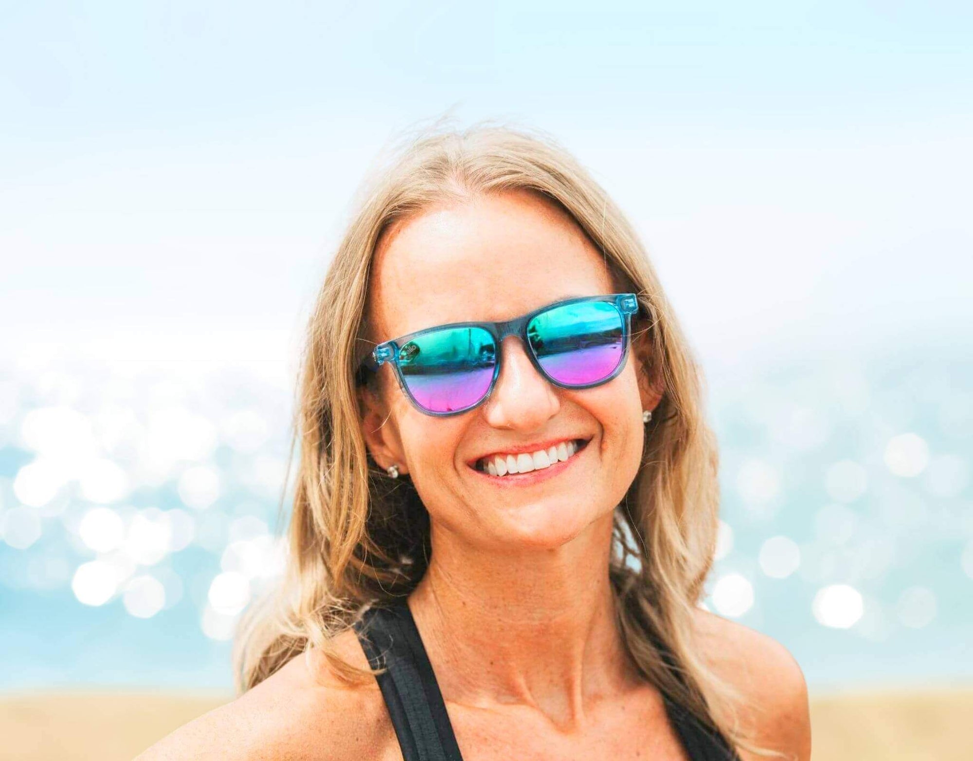 Woman wearing Outer Banks sunglasses for small faces in blue rainbow lens at a sunny beach.