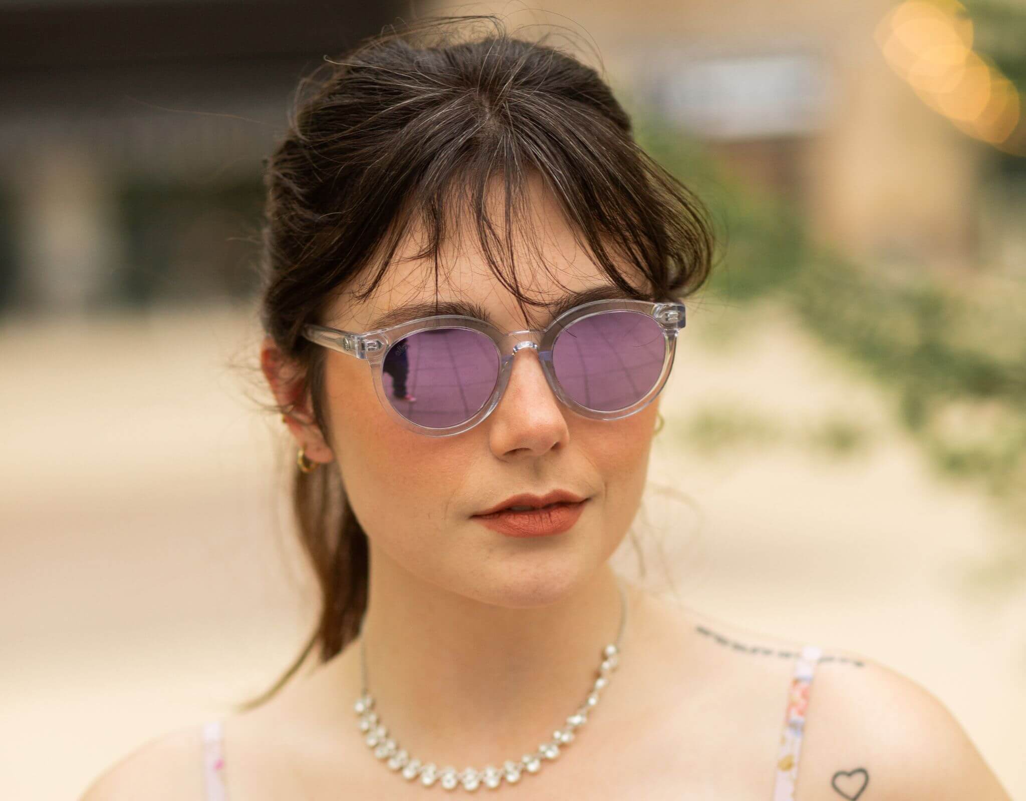 Woman wearing clear purple Bar Harbor small sunglasses for small faces, with soft expression and a blurred outdoor backdrop