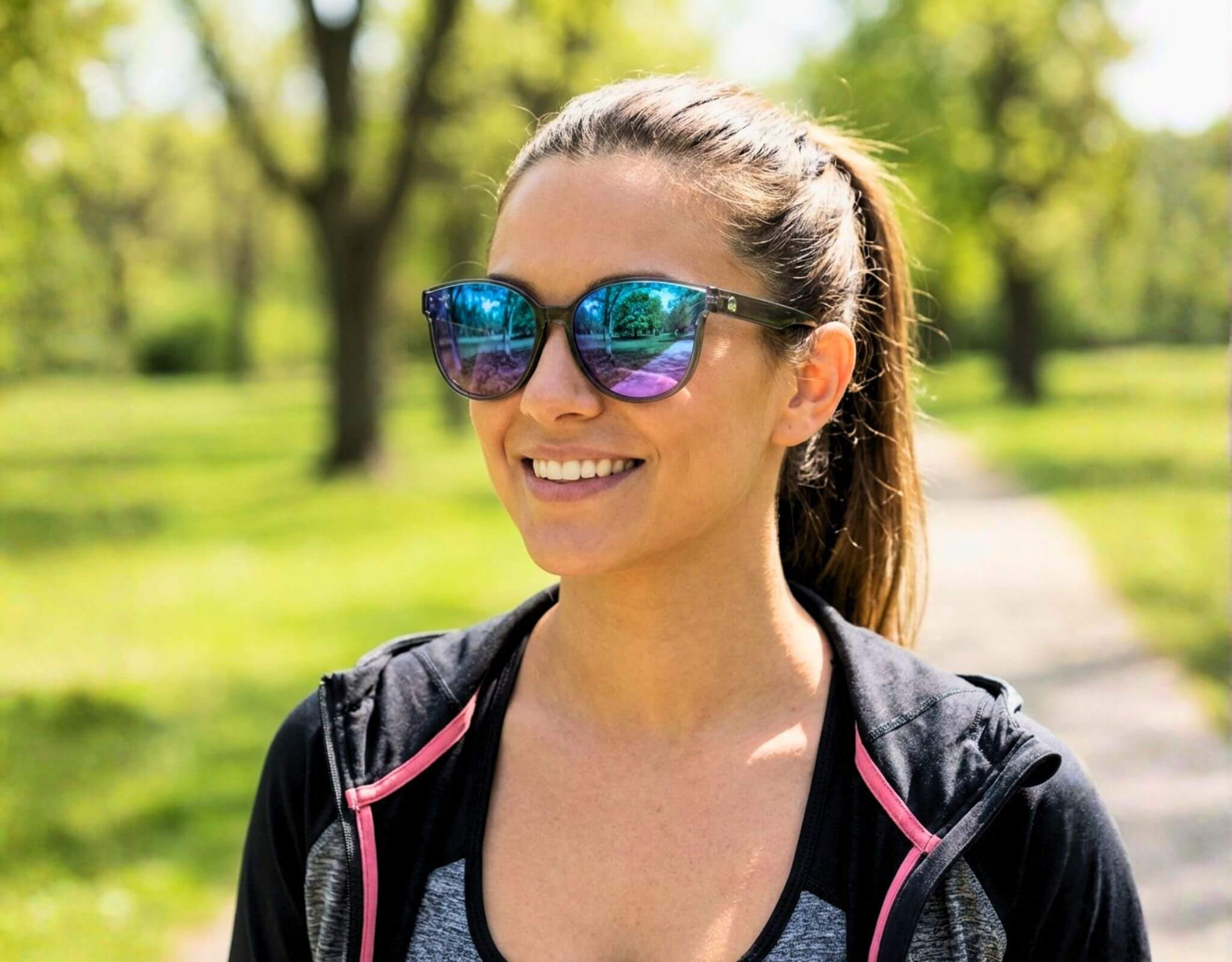 Petite woman with a small head wearing grey and purple South Beach shades for little faces smiling during a walk in a bright green park setting