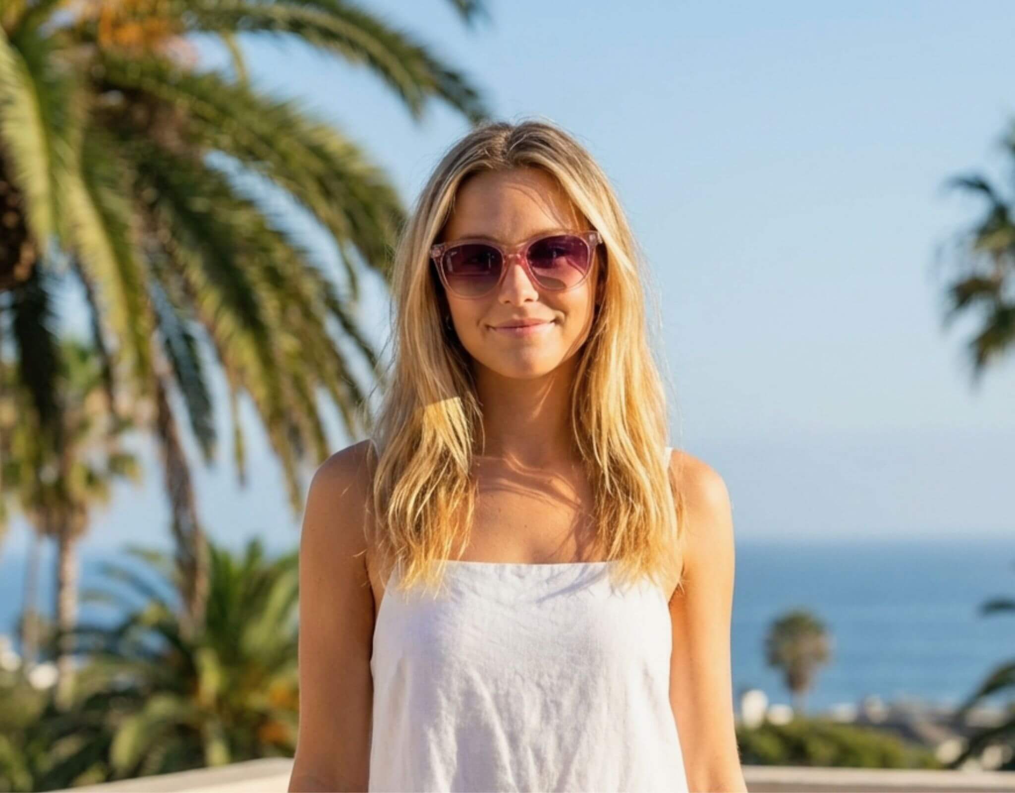 Sunglasses for little faces Pebble Beach in pink frame with lavender lenses worn by a woman with a petite face and small head standing by the ocean in sunny coastal light with ello sunglasses