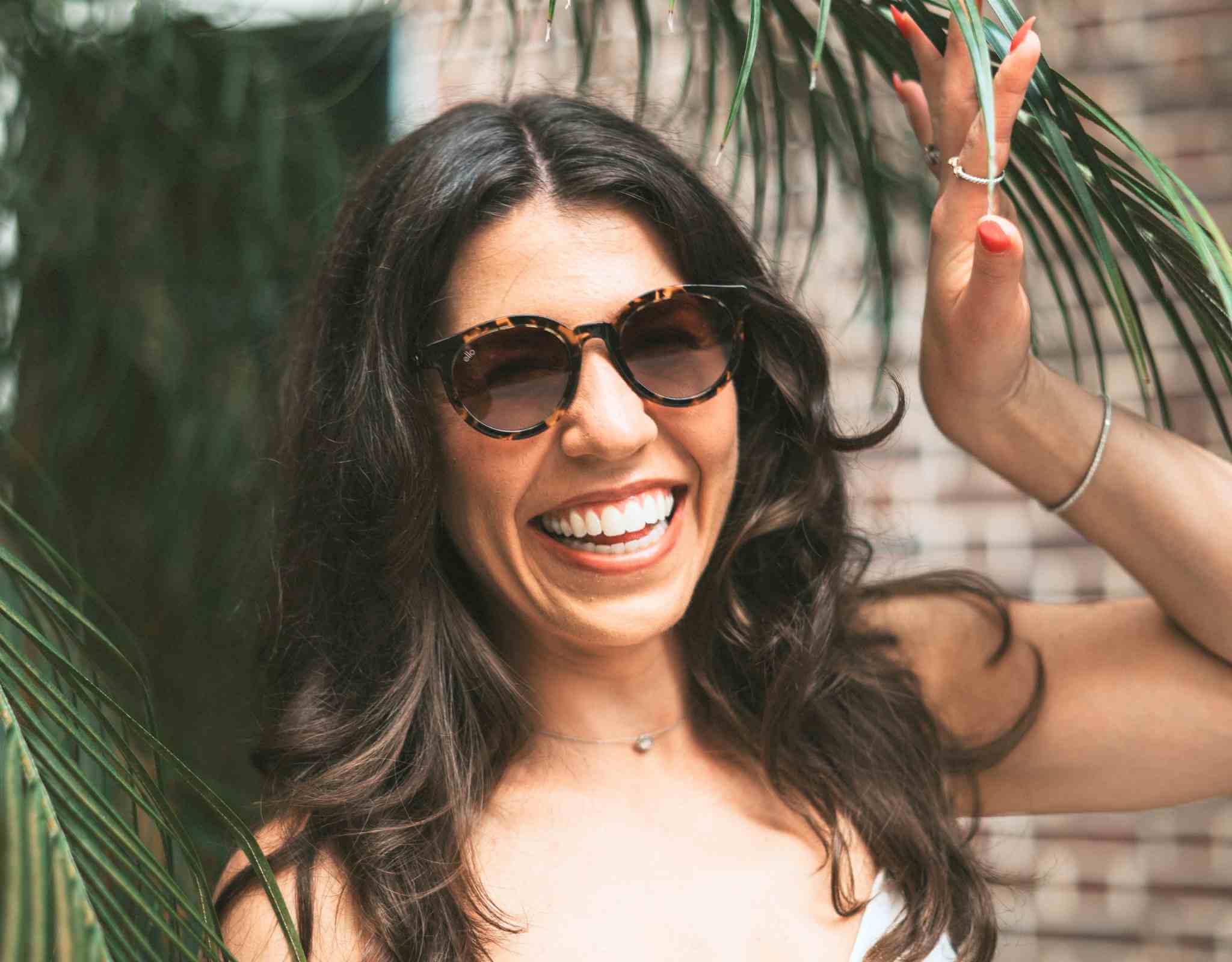Smiling woman wearing tortoise small sunglasses for small faces while standing beside palm leaves.