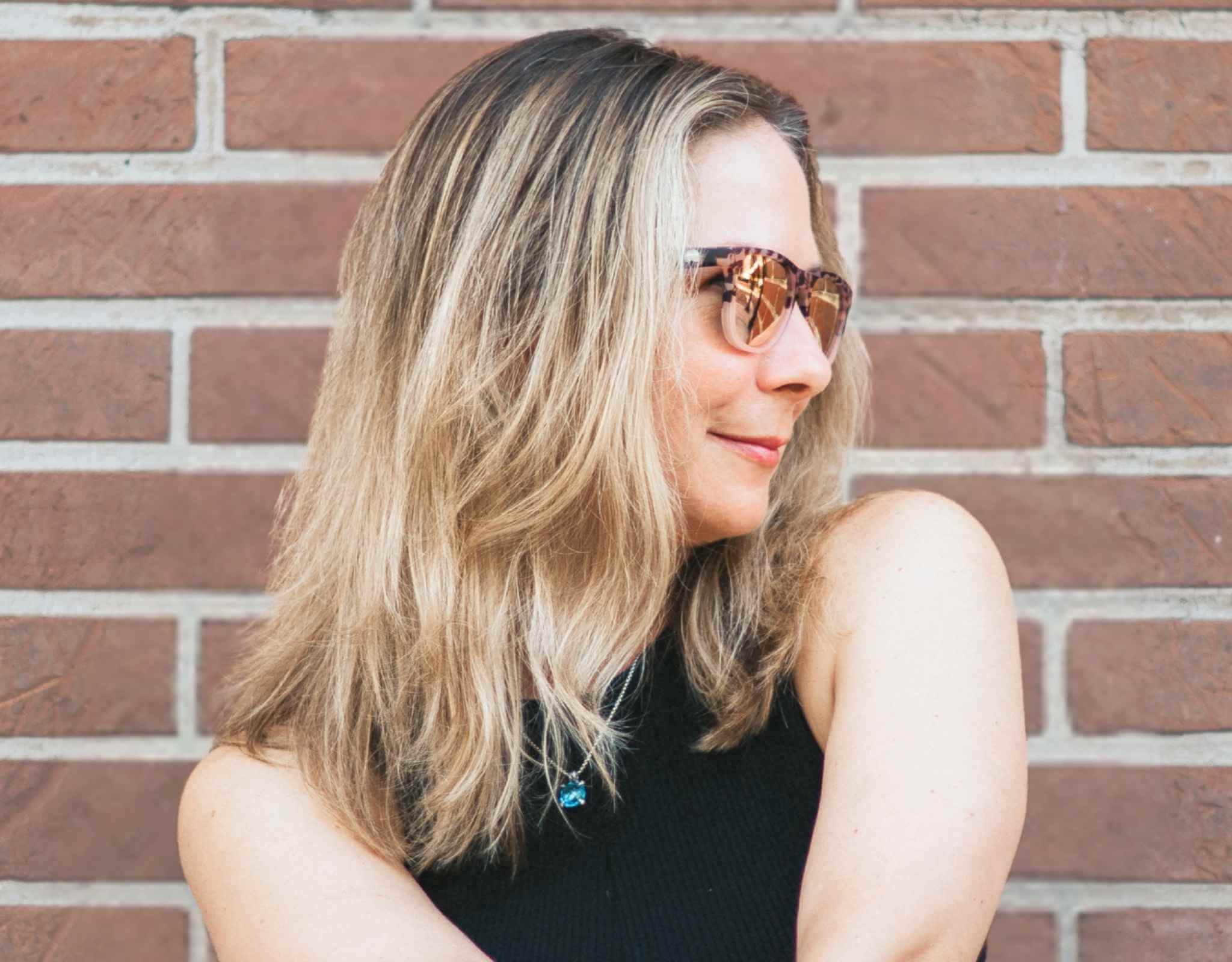 Woman wearing tortoise peach Outer Banks sunglasses for small faces, standing outdoors in front of a red brick wall.